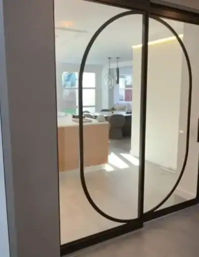 Maatwerk stalen schuifdeur met vast paneel en organisch ovaal design tussen hal en leefruimte.